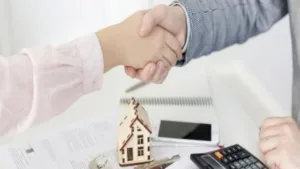 dos personas cerrando un trato con documentos y una maqueta de una casa en la mesa