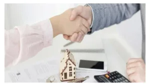 dos personas cerrando un trato con documentos, la maqueta de una casa y una calculadora sobre la mesa